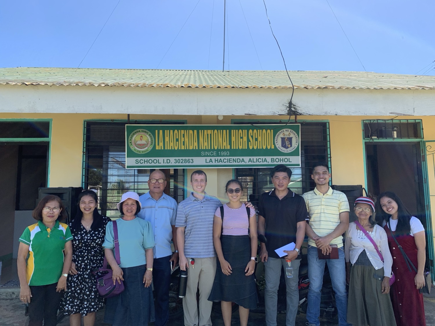 The Schneide family with school staff in front of the La Hacienda National High School sign in Alicia, Bohol